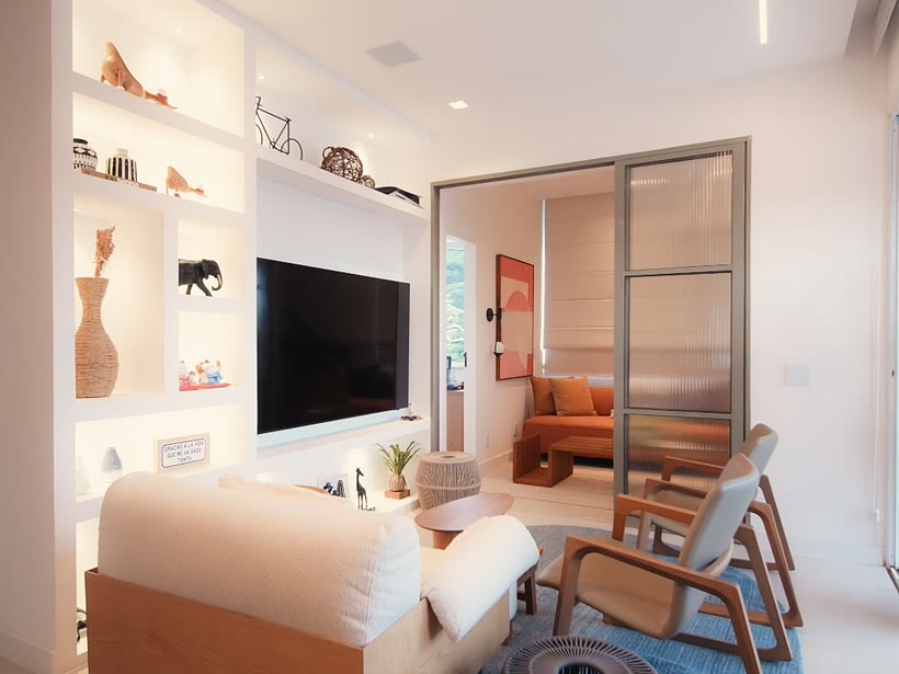 Sala de estar com nichos iluminados, painel de TV e porta de vidro canelado para o quarto